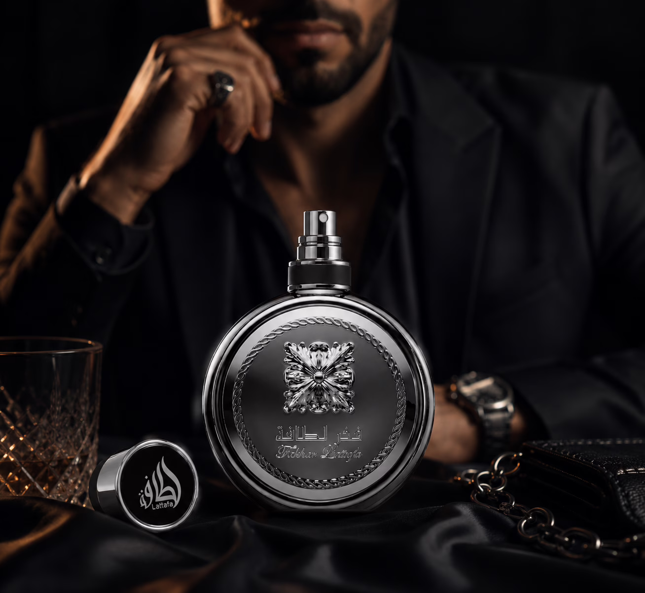 Photographie publicitaire ultra-réaliste du parfum Fakhar Platin de Lattafa, vue de face. Ambiance masculine, sophistiquée et pleine de confiance. Le flacon est au premier plan avec tous ses détails parfaitement nets. En arrière-plan, un homme élégant habillé entièrement en noir se tient dans une posture assurée. Il porte une montre au poignet gauche et une bague à la main droite. Un verre de whisky est placé à gauche du flacon. Éclairage cinématographique, profondeur de champ professionnelle, reflets subtils, atmosphère de luxe moderne, haute définition. Il y a le bouchon ouvert qui est posé à gauche du flacon.
