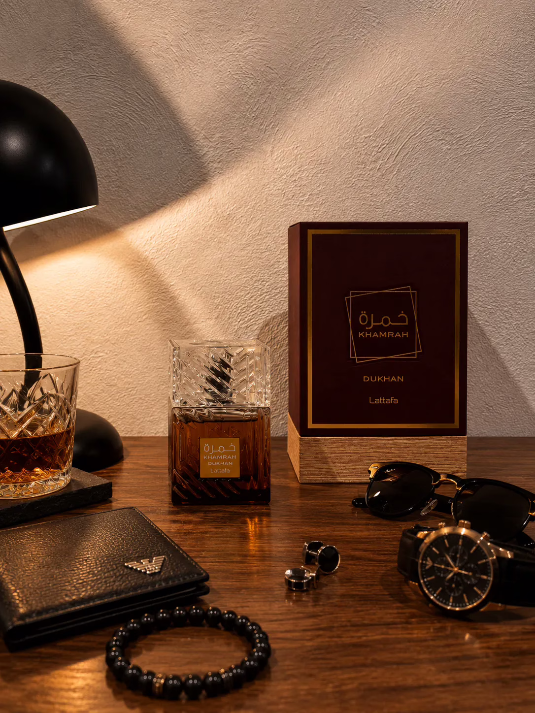 ⁠Parfum Khamrah Dukhan de Lattafa posé sur un bureau en bois avec son coffret, un verre de whisky, une montre noire et un portefeuille.⁠